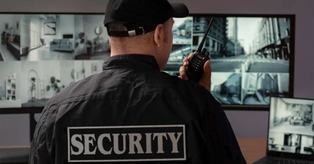 A security guard wearing black jacket sitting in front of cctv screens and speaking on an intercom.