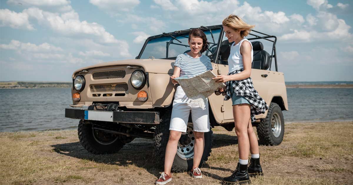 Two beautiful white traveler girls standing and looking at a map while standing in front of a 4x4 truck on a seashore.