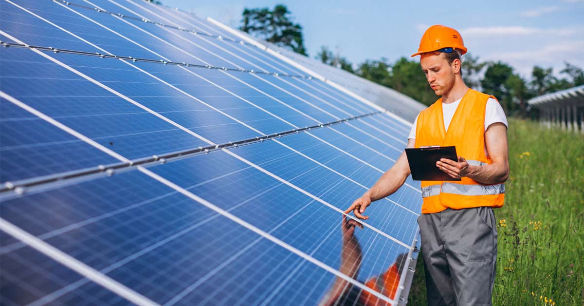 An engineer with orange helmet and orange jacket checking a solar panel with the help of a tablet device.