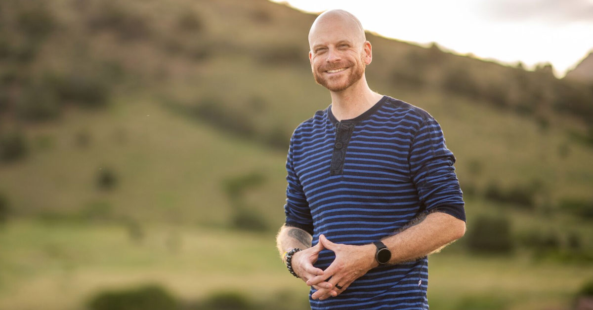 A photo of Nicholas Whitaker in blue hanley t-shirt standing in front of green hills, smiling.