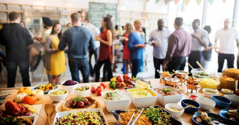 A picture of people networking at an event, with a table full of food items in front.