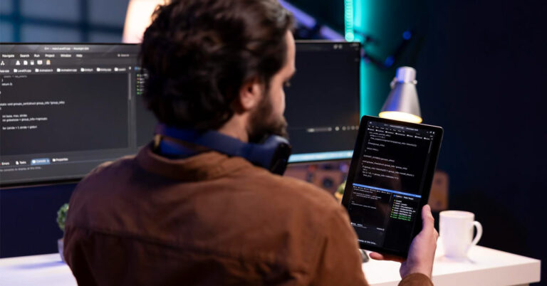 A man testing a software on a desktop and tablet. the picture is clicked from behind. the guys is holding the tablet in his right hand