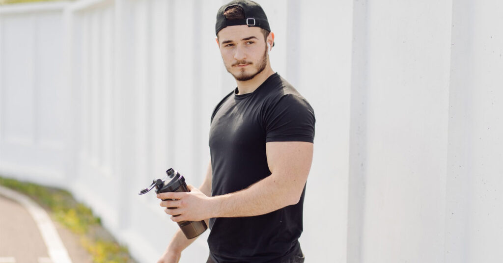 A white guy wearing black half-sleeves T-shirt holding a sipper in hand and wearing a black cap