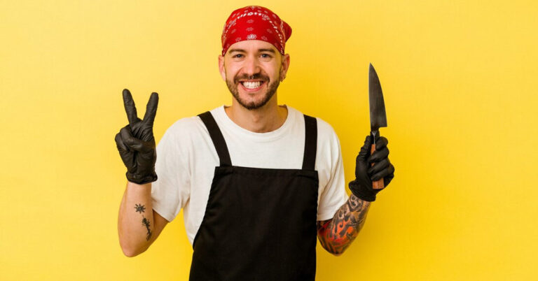 A modern male butcher standing in front of yellow background holding a knife in left hand and making peace symbol from the right hand