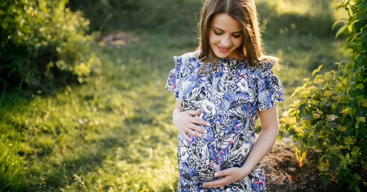 A pregnant blonde white woman wearing a floral dress - standing in a garden