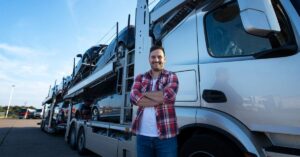 A male vehicle transporter standing with hands folded in front of a large vehicle transportation truck