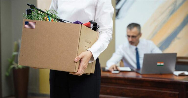 A female employee holding a cardboard box - leaving the office. The male boss sitting on the desk in the background