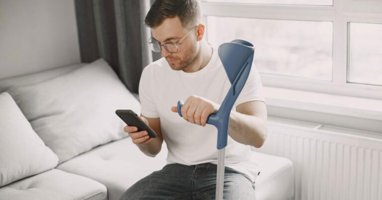 A young white man sitting, holding a crutch in left hand, and operating a smartphone with right hand.