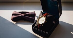 A leather strap gift watch in case kept on a table along with a bow-tie