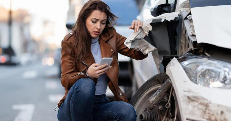 A caucasian woman with sad expression sitting by her damaged car front and operating her smartphone.