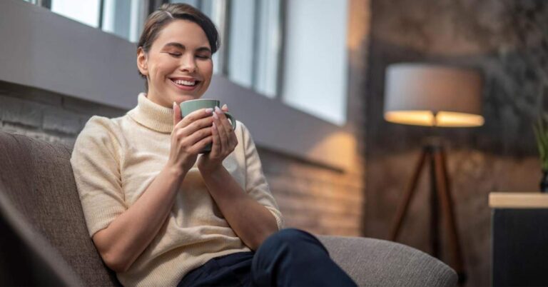 A white women sitting and enjoying her cup of coffee on a sofa.