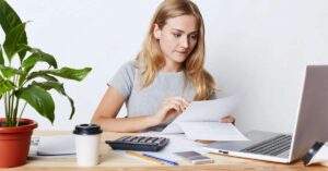 A girl holding papers, watching laptop screen, a calculator is kept along