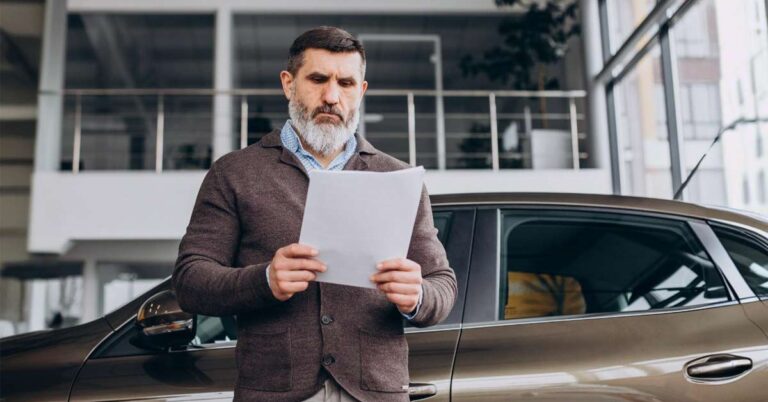 A middle-aged handsome man wearing a waistcoat standing and reading car document papers in front of a car.