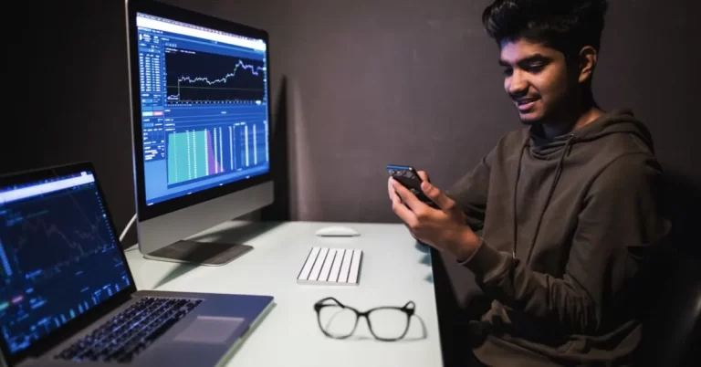 A young south asian man with happy expression operating smartphone while sitting in front of a computer screen depicting a trading graph.