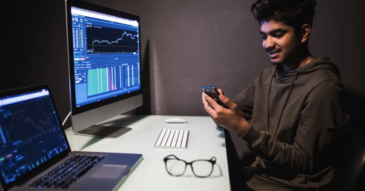 A young south asian man with happy expression operating smartphone while sitting in front of a computer screen depicting a trading graph.