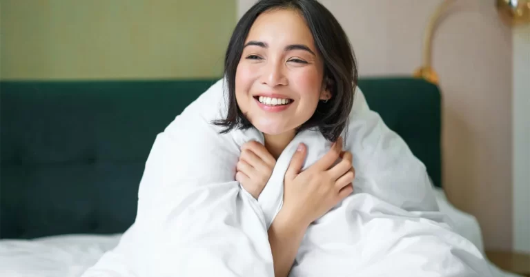 An asian woman with a happy face sitting wrapped in a white quilt.