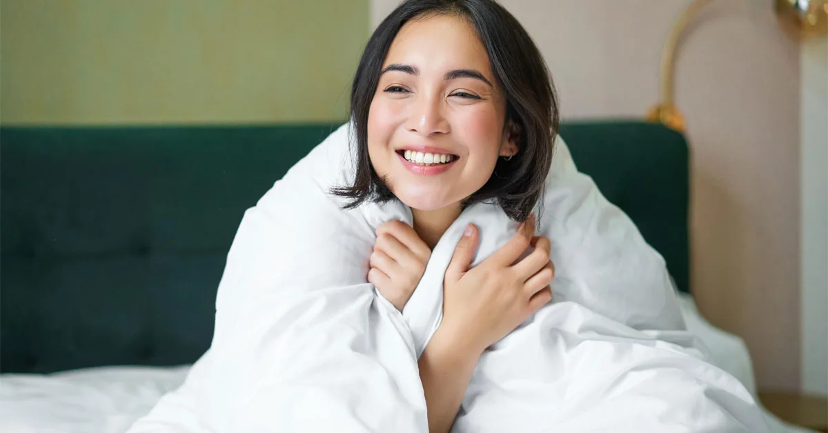 An asian woman with a happy face sitting wrapped in a white quilt.