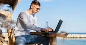 A young man working on a laptop while traveling on a beach - depicting a digital nomad concept