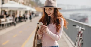 Young woman traveler standing on a waterfront path, looking at her smartphone while wearing a hat and earbuds, illustrating phone safety and travel tips.