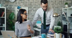 A young engineer at a laptop looking detached while an older manager stands over the desk in a startup office