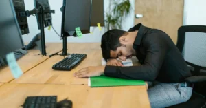 An exhausted man in a black shirt rests his head on his desk at an office, surrounded by monitors and documents