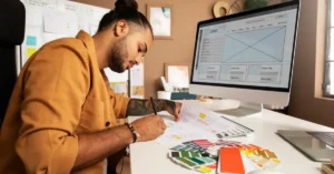 A designer sketching wireframes by hand with colour swatches on the desk and a website layout on the monitor behind