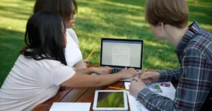 Three students gathered around a laptop reviewing a shared Word document, reflecting how group coursework now happens through collaborative digital tools rather than in-person meetups.