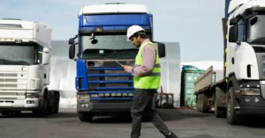 Worker in a white hard hat and hi-vis vest walking past parked trucks in a depot yard