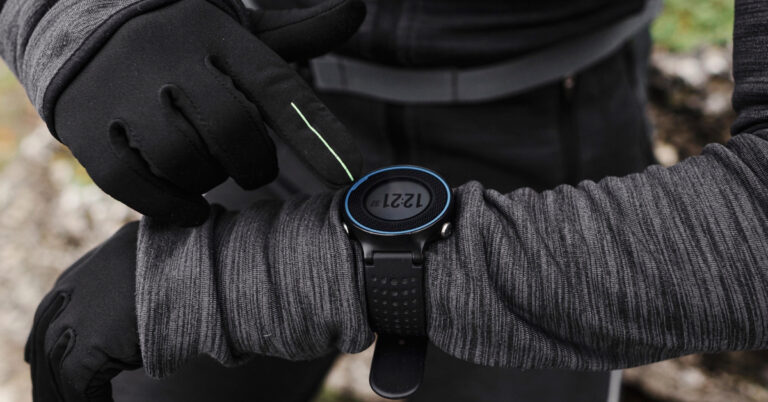 Hiker in gloves tapping a black digital sports watch with a blue bezel on a rocky trail